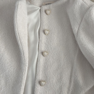 Könnte beinhalten: Cremefarbene Jacke mit herzförmigen Knöpfen. Die Jacke hat ein strukturiertes Gewebe und ein Satinfutter. Die goldfarbenen Knöpfe sind mit kristallartigen Verzierungen versehen. Ein klassischer Stil, geeignet für verschiedene Anlässe.