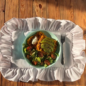 Könnte beinhalten: Ein hellblauer Teller mit Waffeln, Avocado und Salat steht auf einer weißen Tischdecke mit Rüschenkante. Eine silberne Gabel liegt daneben. Das Essen steht auf einem Holztisch.