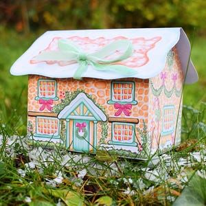 May include: A house-shaped gift box featuring a printed orange stone texture, a light blue roof with a pale green ribbon bow, and illustrations of windows with pink bows, a green wreath on the front door, and festive garlands on the exterior walls.