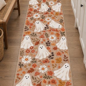 May include: A long rectangular runner rug featuring a repeating pattern of white ghosts, orange and red autumn leaves, small red mushrooms with white spots, and white daisy flowers on a light tan background with a finished bound edge.