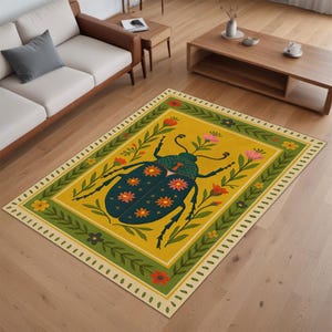 Op de afbeelding: Een rechthoekig vloerkleed met een grote, donkerblauwe kever in het midden op een mosterdgele achtergrond. De kever is versierd met oranje en roze bloemmotieven. Een groene rand met blad- en bloemmotieven omlijst het centrale ontwerp.