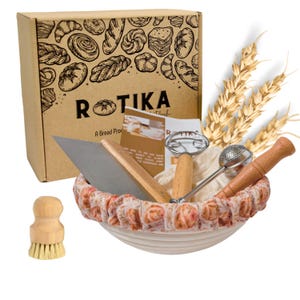 May include: A set of bread baking tools in a white proofing basket with a floral pattern. The set includes a dough scraper, a proofing basket liner, a bread lame, a dough whisk, a bread scoring tool, and a brush. The tools are arranged in the basket with a box of bread baking supplies in the background.