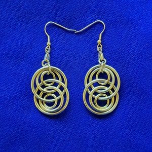 Puede incluir: Un par de pendientes dorados con un diseño intrincado e entrelazado. Cada pendiente presenta una serie de bucles entrelazados y un gancho simple. Los pendientes se muestran sobre un fondo azul vibrante.