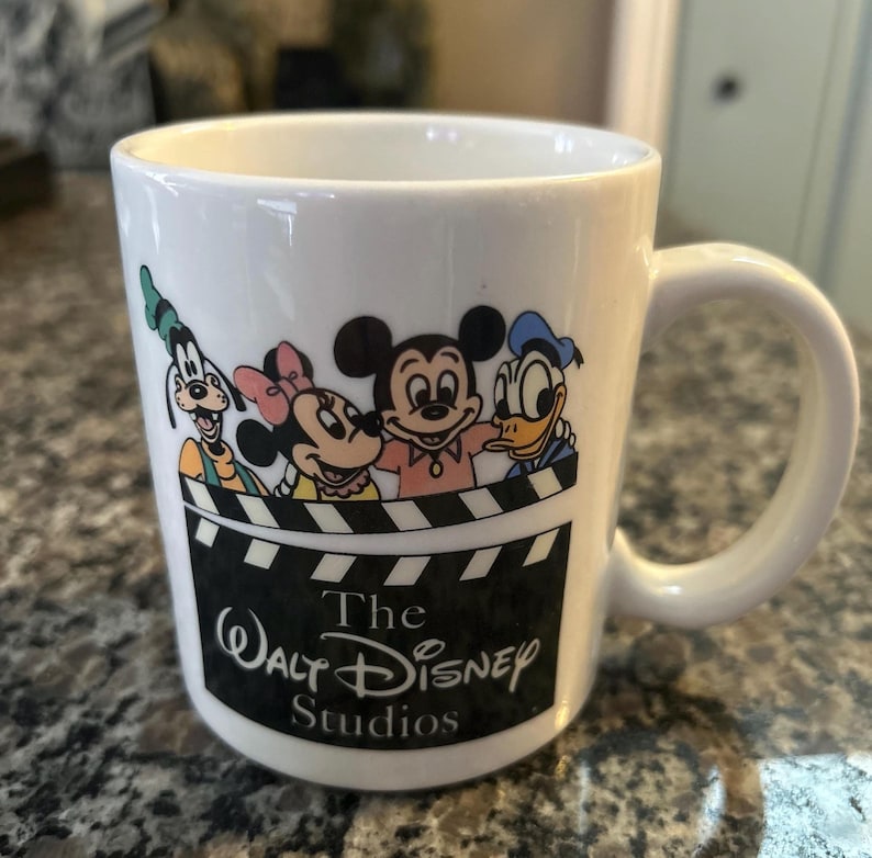 May include: A white ceramic mug featuring a graphic of Goofy, Minnie Mouse, Mickey Mouse, and Donald Duck peering over a black and white film clapperboard. The text "The Walt Disney Studios" is printed in white on the black clapperboard section.