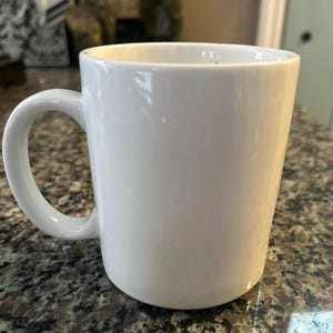 May include: A plain white ceramic mug with a smooth, glossy finish and a curved handle on the left side. The mug features a simple, cylindrical body with a wide opening at the top and a solid base, presenting a clean and minimalist appearance.