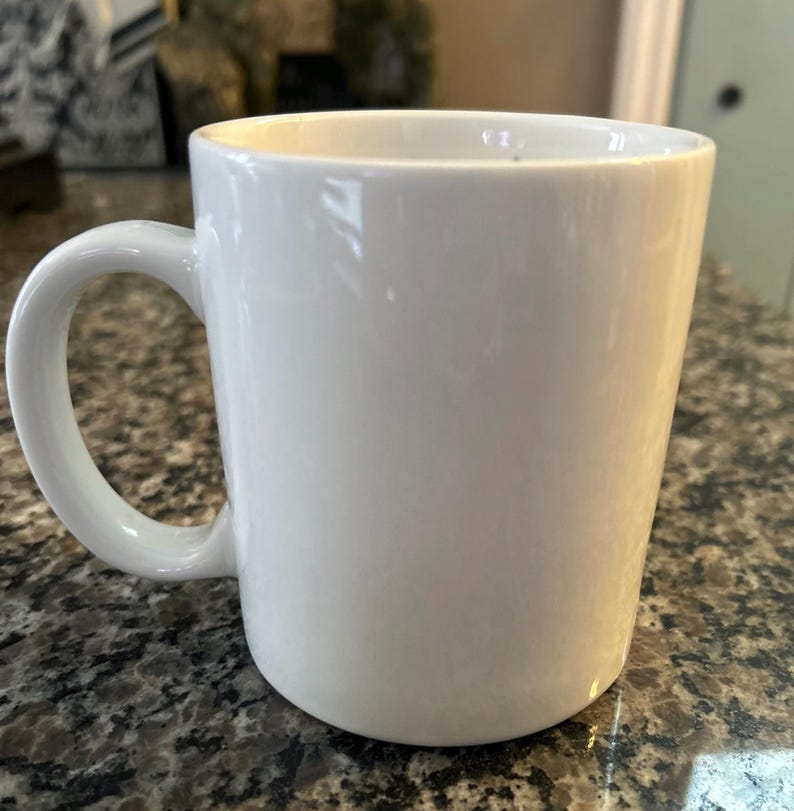 May include: A plain white ceramic mug with a smooth, glossy finish and a curved handle on the left side. The mug features a simple, cylindrical body with a wide opening at the top and a solid base, presenting a clean and minimalist appearance.