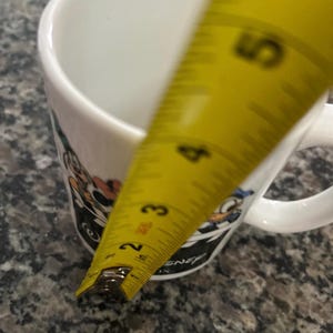 May include: A white ceramic mug featuring colorful cartoon character graphics and the text Disney. A yellow measuring tape is positioned vertically against the side of the mug, showing a height measurement of approximately 4.5 inches from the base.