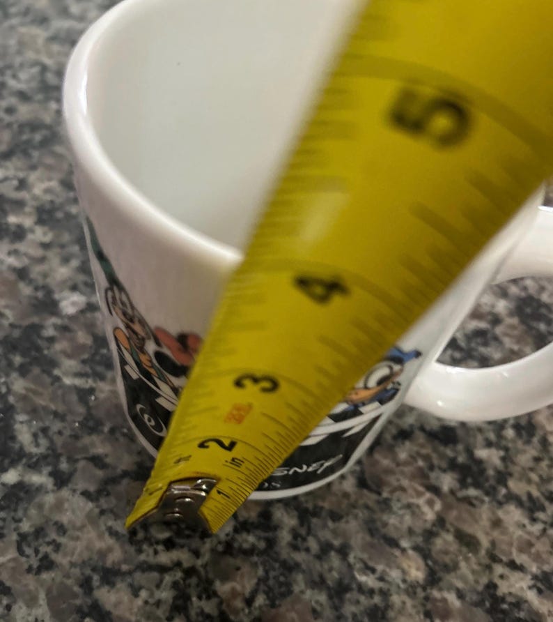 May include: A white ceramic mug featuring colorful cartoon character graphics and the text Disney. A yellow measuring tape is positioned vertically against the side of the mug, showing a height measurement of approximately 4.5 inches from the base.
