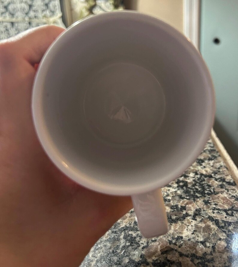 May include: A white ceramic mug viewed from directly above, showing the circular opening and the interior base. The mug features a smooth, glossy white finish on both the interior and exterior surfaces, with a single handle extending from the side.