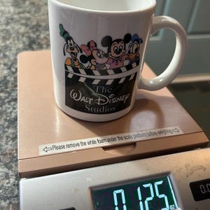 May include: A white ceramic mug featuring a graphic of Goofy, Minnie Mouse, Mickey Mouse, and Donald Duck above a film clapperboard design with the text The Walt Disney Studios. The mug sits on a digital scale displaying a weight of 0.125 lb.