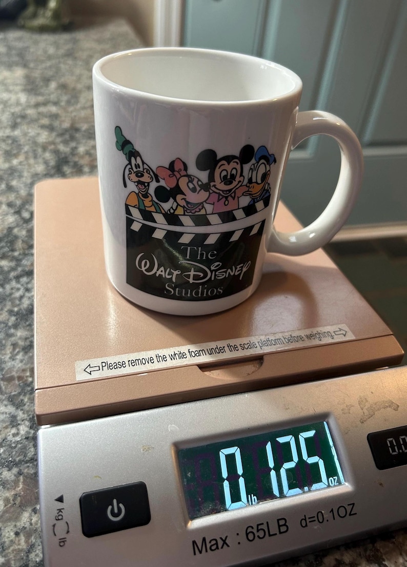 May include: A white ceramic mug featuring a graphic of Goofy, Minnie Mouse, Mickey Mouse, and Donald Duck above a film clapperboard design with the text The Walt Disney Studios. The mug sits on a digital scale displaying a weight of 0.125 lb.