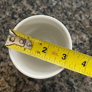 May include: A white ceramic cup viewed from above, positioned next to a yellow measuring tape that shows the opening of the cup has a diameter of approximately 2.5 inches. The cup has a smooth, plain interior and a simple circular rim.