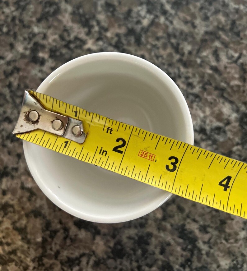 May include: A white ceramic cup viewed from above, positioned next to a yellow measuring tape that shows the opening of the cup has a diameter of approximately 2.5 inches. The cup has a smooth, plain interior and a simple circular rim.