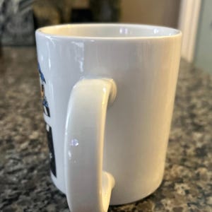 May include: A white ceramic mug with a curved handle on the side. The exterior surface features a partial view of a graphic design with blue and black elements visible on the left side of the mug, while the rest of the mug is plain white.