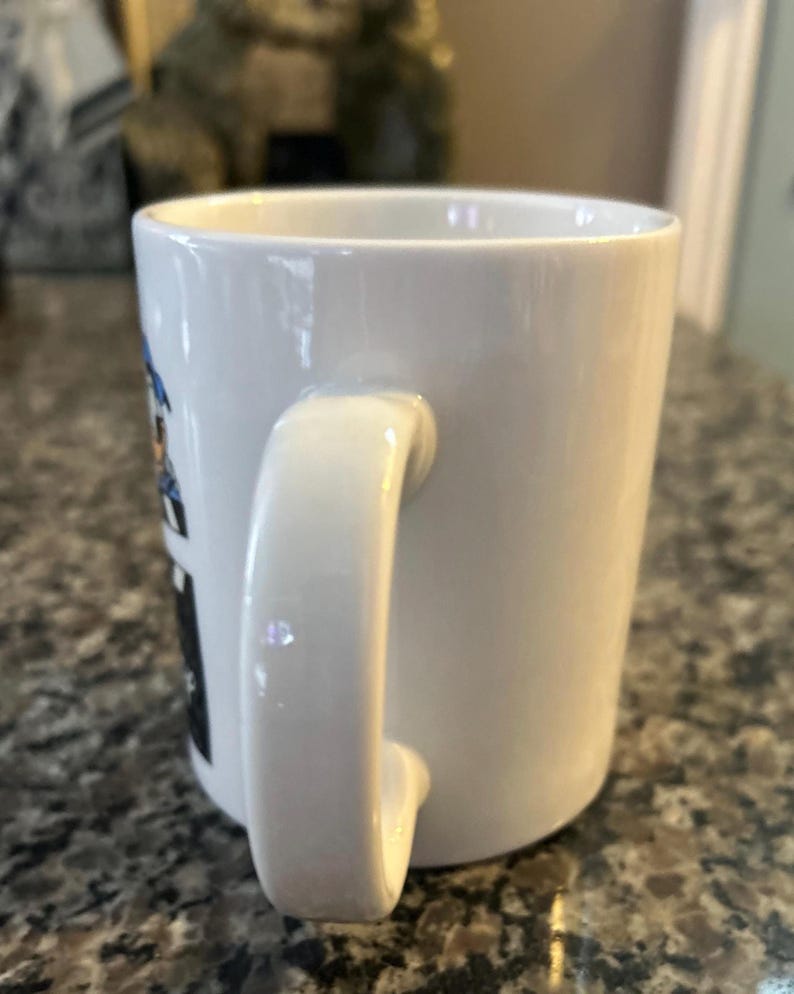 May include: A white ceramic mug with a curved handle on the side. The exterior surface features a partial view of a graphic design with blue and black elements visible on the left side of the mug, while the rest of the mug is plain white.