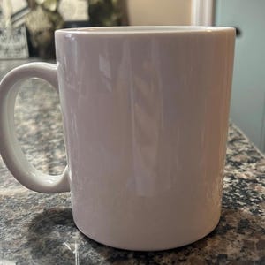 May include: A plain white ceramic mug with a smooth, glossy finish and a curved handle on the left side. The mug features a cylindrical body with a flat base and a wide, open top, presenting a clean, minimalist design suitable for hot beverages.