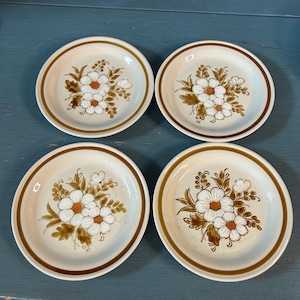 Może przedstawiać: Cztery talerze ceramiczne w stylu vintage z kwiatowym wzorem. Każdy talerz ma biały kwiat z brązowymi i pomarańczowymi akcentami, otoczony brązowymi liśćmi i brązową obwódką. Talerze są ustawione na niebieskim tle.
