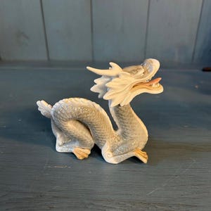 May include: A porcelain dragon figurine featuring a coiled body with textured scales in shades of light gray and white. The creature has a detailed head with prominent horns, an open mouth revealing a pink tongue, and small tan-colored claws.