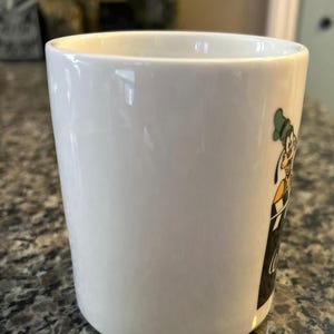 May include: A white ceramic mug featuring a graphic of the cartoon character Goofy wearing a green hat and yellow shirt, positioned above a black rectangular area with white cursive text that appears to be partially visible on the side.