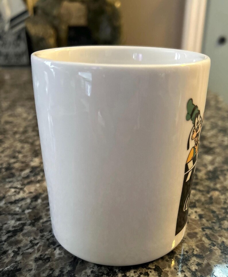 May include: A white ceramic mug featuring a graphic of the cartoon character Goofy wearing a green hat and yellow shirt, positioned above a black rectangular area with white cursive text that appears to be partially visible on the side.
