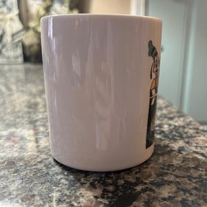 May include: A white ceramic mug featuring a graphic of the Disney character Goofy wearing a green hat and orange shirt, positioned behind a black and white film clapperboard. The mug has a smooth, glossy finish and a cylindrical shape.