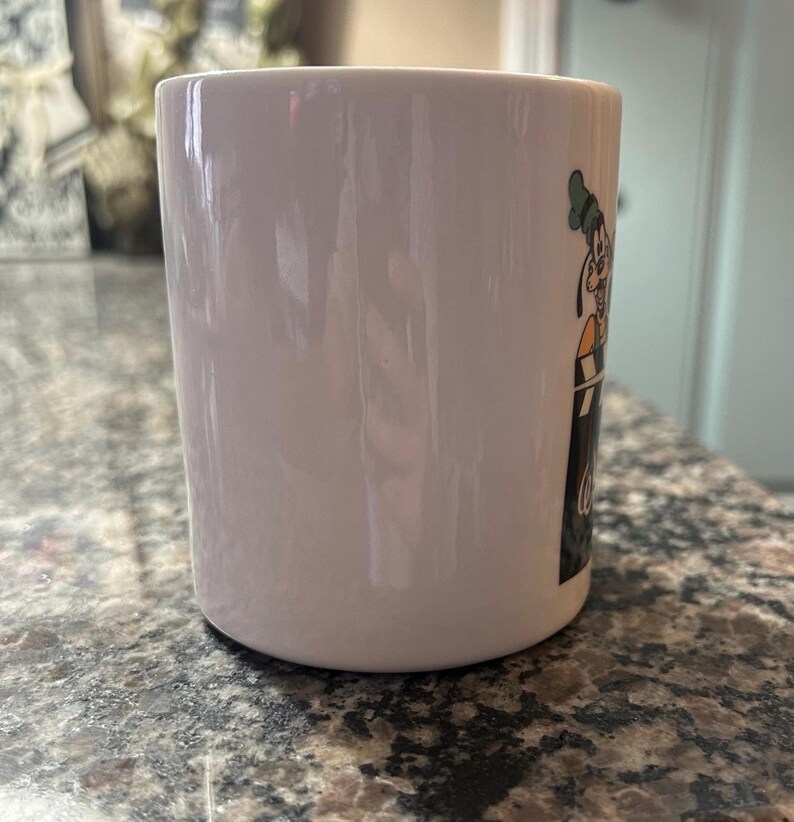 May include: A white ceramic mug featuring a graphic of the Disney character Goofy wearing a green hat and orange shirt, positioned behind a black and white film clapperboard. The mug has a smooth, glossy finish and a cylindrical shape.