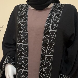 May include: Black open-front abaya with white geometric embroidery along the edges and sleeves. The abaya is worn over a taupe dress and a black hijab. The design features a modern, elegant aesthetic.
