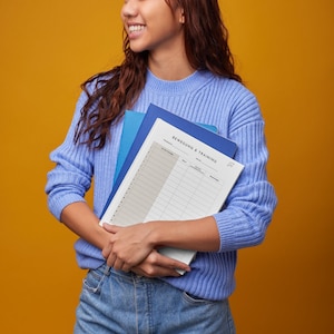 May include: A woman holding a blue and white planner with the text "Bewegung & Training" on the cover. The planner has a grid layout with columns for dates, activities, and notes.