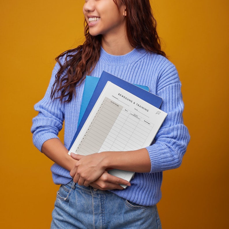 May include: A woman holding a blue and white planner with the text "Bewegung & Training" on the cover. The planner has a grid layout with columns for dates, activities, and notes.