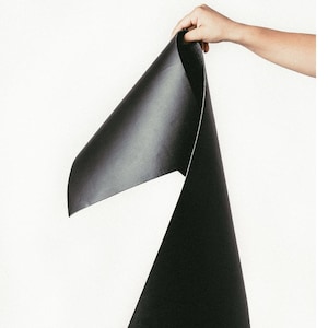 May include: A hand holds up a large, black, textured sheet of material against a white background. The material is folded, revealing a lighter underside. The image highlights the texture and color of the material.