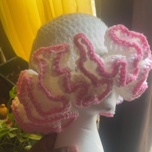 May include: A white crocheted hat with a pink ruffled trim. The hat is displayed on a white mannequin head. The hat's texture is visible, and the pink trim adds a pop of color. The hat is a handmade item.