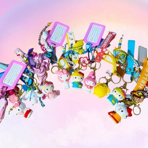 Può includere: Un set di portachiavi colorati con personaggi dei cartoni animati, tra cui Hello Kitty, con accenti rosa, gialli, blu e rossi. I portachiavi sono attaccati a porta tessere d'identità rosa e gialli con il testo "Buona fortuna" e "Ciao".