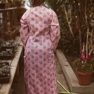 May include: A long, pink floral-print dress with long sleeves. The dress features a repeating pattern of small blue and yellow flowers. The person wearing the dress is standing in a greenhouse, surrounded by plants and flowers.