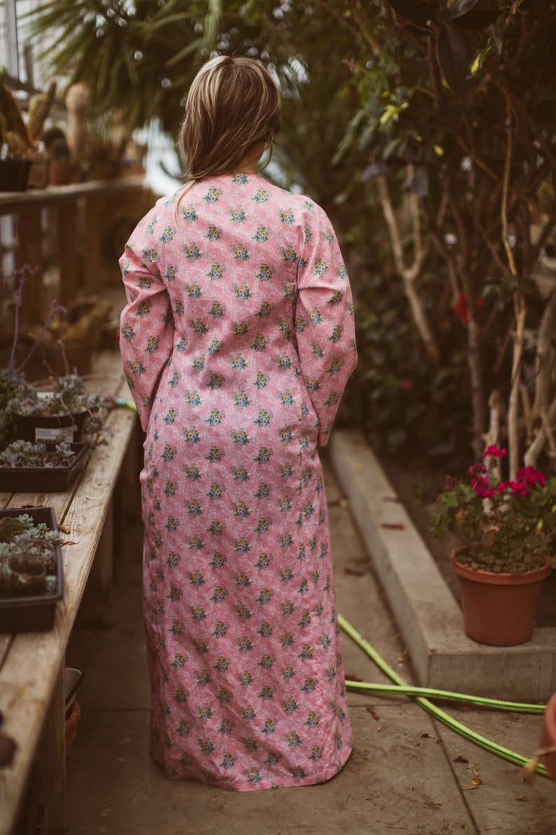 May include: A long, pink floral-print dress with long sleeves. The dress features a repeating pattern of small blue and yellow flowers. The person wearing the dress is standing in a greenhouse, surrounded by plants and flowers.