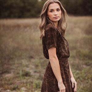 Puede incluir: Una mujer con un vestido marr&oacute;n de manga corta con estampado de leopardo. El vestido tiene cintura entallada y falda acampanada. Est&aacute; de pie en un campo con hierba alta y &aacute;rboles al fondo.