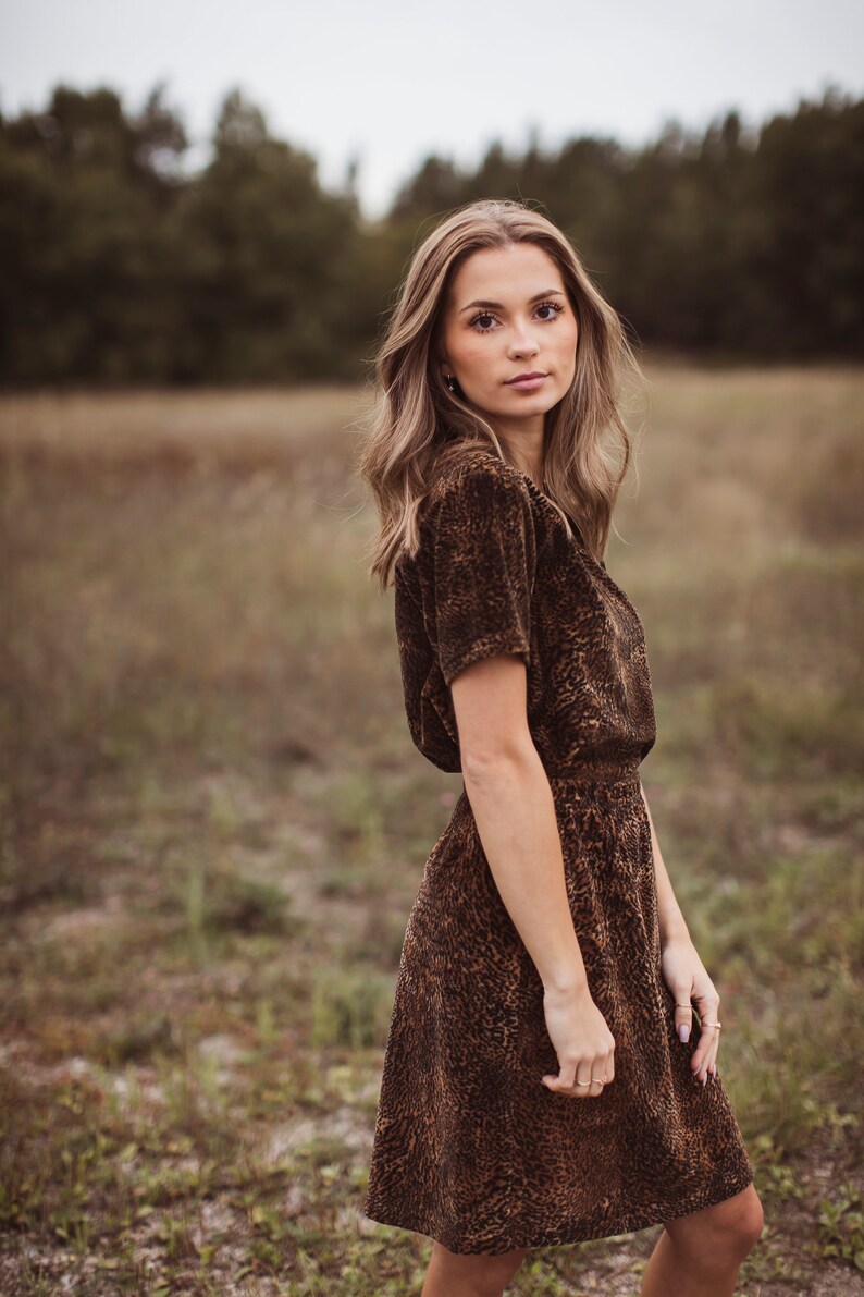Puede incluir: Una mujer con un vestido marr&oacute;n de manga corta con estampado de leopardo. El vestido tiene cintura entallada y falda acampanada. Est&aacute; de pie en un campo con hierba alta y &aacute;rboles al fondo.
