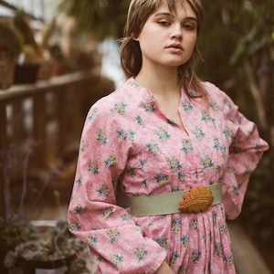 May include: A pink floral dress with long sleeves and a mandarin collar. The dress is cinched at the waist with a light green belt and a brown leaf-shaped buckle. The background is a greenhouse setting.