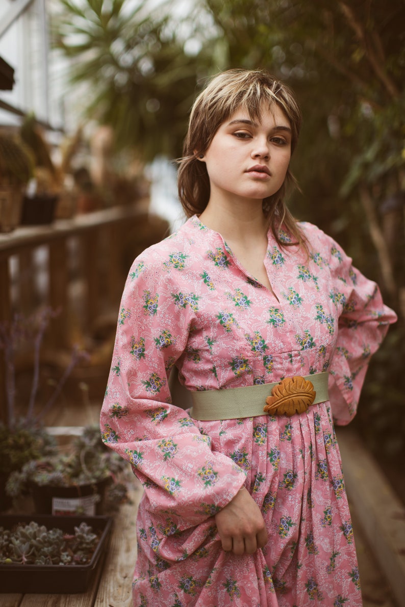 May include: A pink floral dress with long sleeves and a mandarin collar. The dress is cinched at the waist with a light green belt and a brown leaf-shaped buckle. The background is a greenhouse setting.