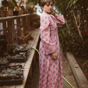 May include: A pink floral print dress with long sleeves. The dress is knee-length and features a delicate pattern of small blue and yellow flowers. The image is taken in a greenhouse setting, with plants and wooden shelves in the background.