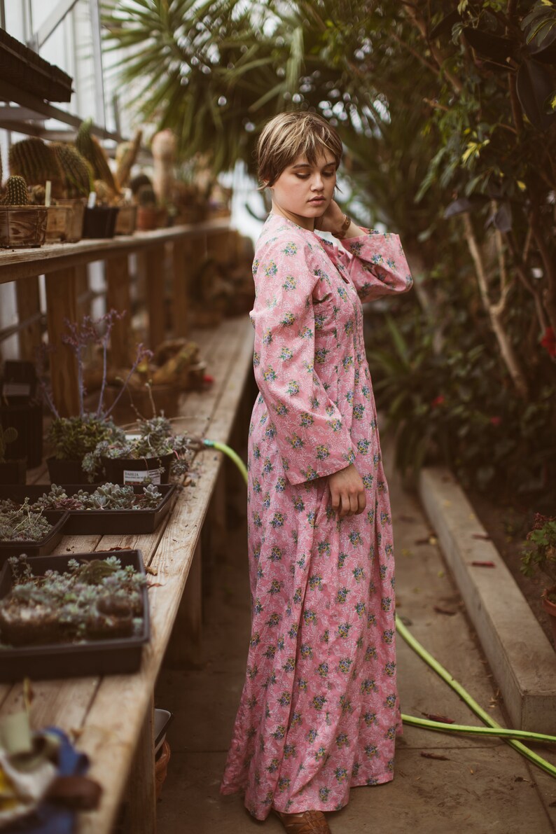 May include: A pink floral print dress with long sleeves. The dress is knee-length and features a delicate pattern of small blue and yellow flowers. The image is taken in a greenhouse setting, with plants and wooden shelves in the background.