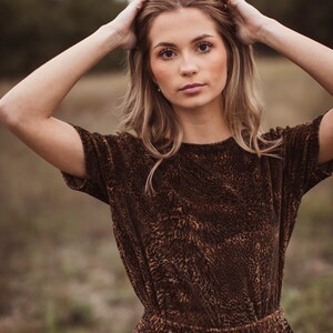 Puede incluir: Una mujer con un vestido marr&oacute;n de manga corta con estampado de leopardo. El vestido tiene la cintura ajustada y la mujer se lleva las manos a la cabeza. El fondo es una escena borrosa al aire libre.