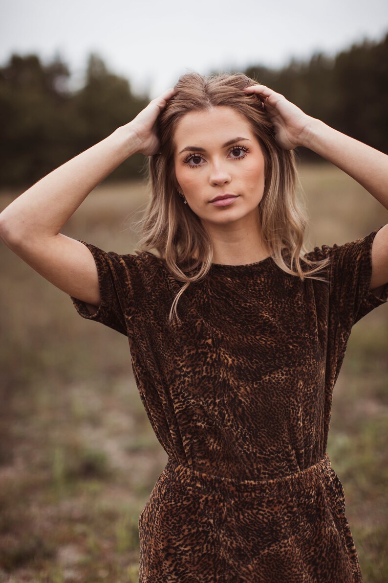 Puede incluir: Una mujer con un vestido marr&oacute;n de manga corta con estampado de leopardo. El vestido tiene la cintura ajustada y la mujer se lleva las manos a la cabeza. El fondo es una escena borrosa al aire libre.