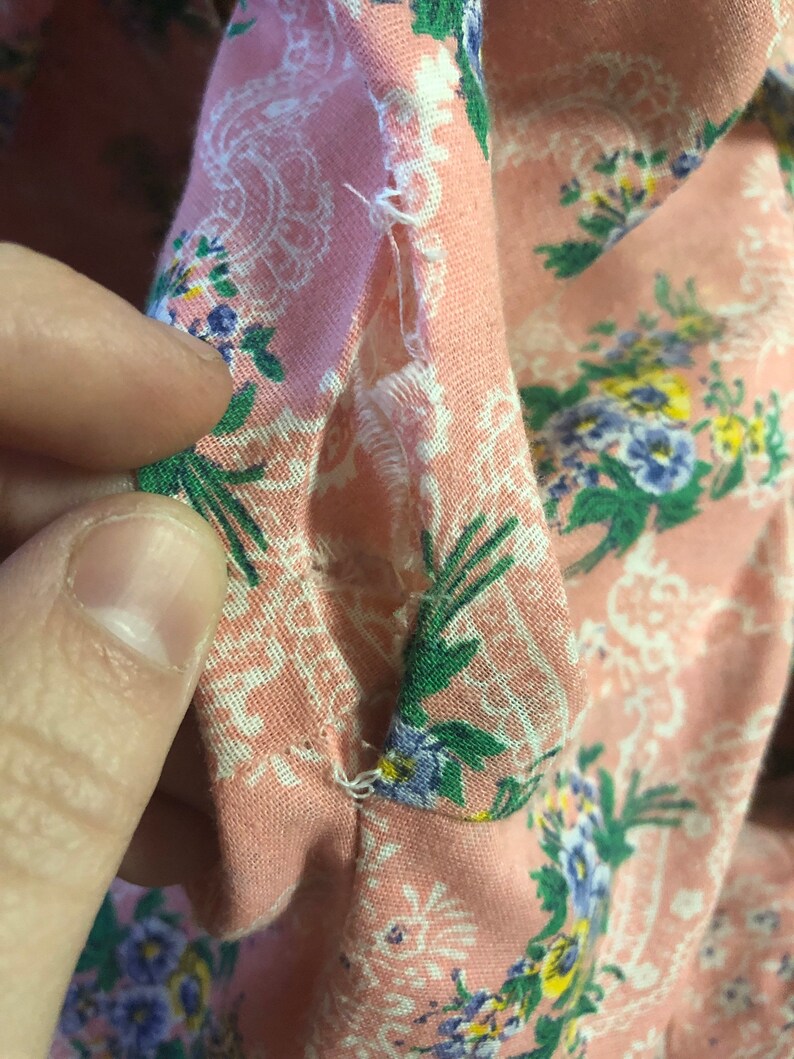 May include: Close-up of a section of pink floral fabric with a white paisley design. The fabric has a tear in the seam, revealing the raw edges and stitching. The fabric is decorated with small blue, green, and yellow flowers.
