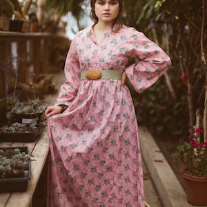 May include: A pink floral print dress with long sleeves and a wide, light green belt. The dress features a leaf-shaped buckle and a v-neckline. The model is standing in a greenhouse setting with plants in the background.