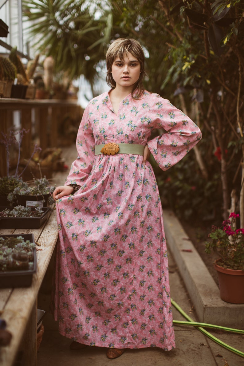 May include: A pink floral print dress with long sleeves and a wide, light green belt. The dress features a leaf-shaped buckle and a v-neckline. The model is standing in a greenhouse setting with plants in the background.