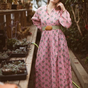 May include: A pink floral print dress with long sleeves and a V-neck. The dress is cinched at the waist with a light green belt and a wooden leaf-shaped buckle. The background features a greenhouse setting with plants.