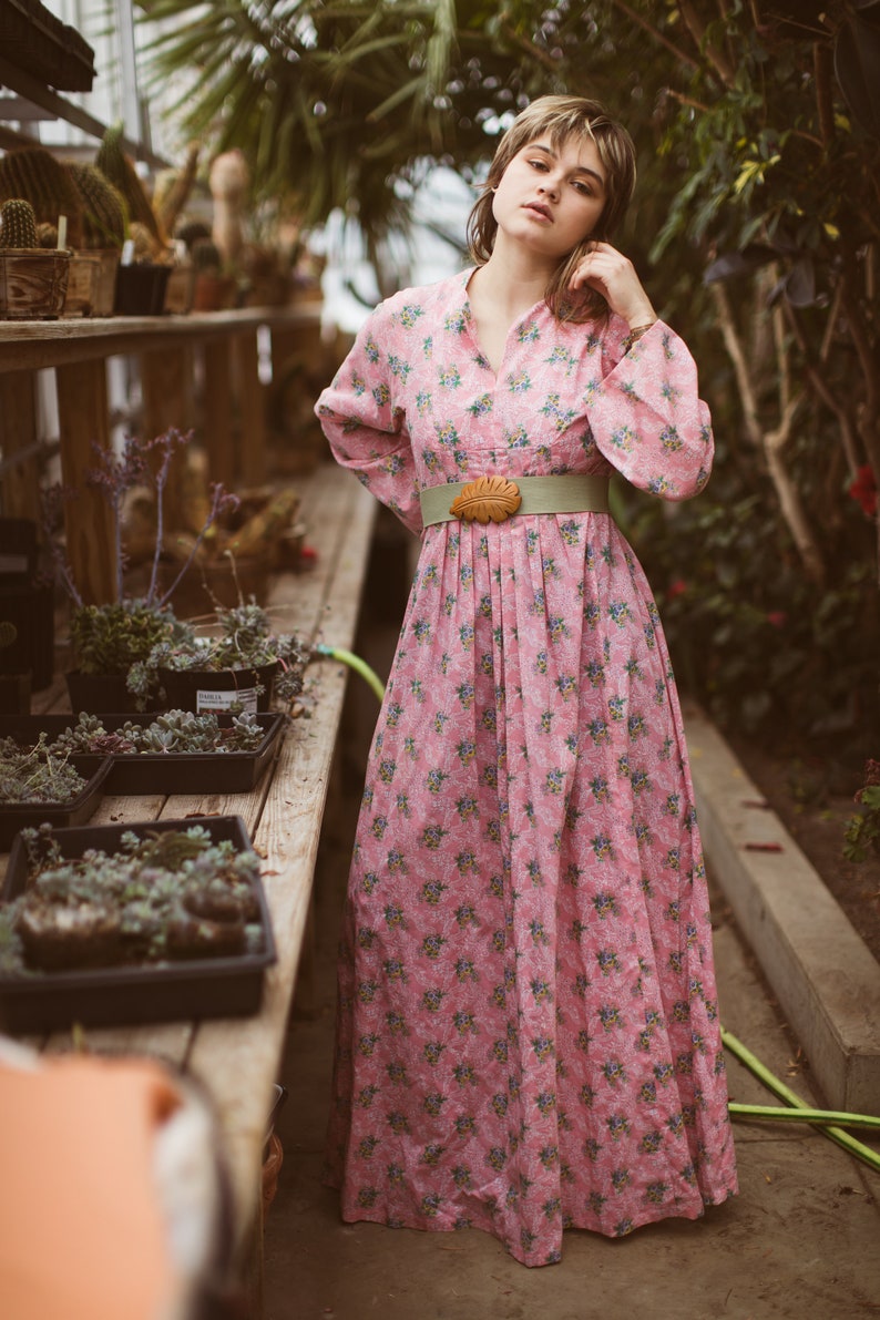 May include: A pink floral print dress with long sleeves and a V-neck. The dress is cinched at the waist with a light green belt and a wooden leaf-shaped buckle. The background features a greenhouse setting with plants.