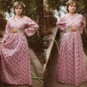 May include: A long, pink floral dress with wide sleeves and a v-neck. The dress is cinched at the waist with a light green belt and a wooden buckle. The dress has a floral pattern with green and blue accents.