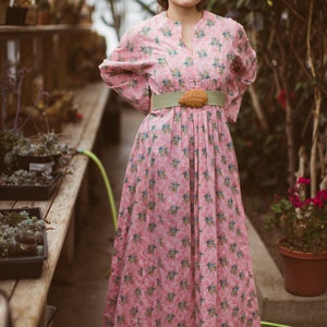 May include: A long pink dress with a floral pattern, featuring a high neckline and long sleeves. The dress is cinched at the waist with a light green belt and a decorative buckle. The dress is worn in a greenhouse setting.