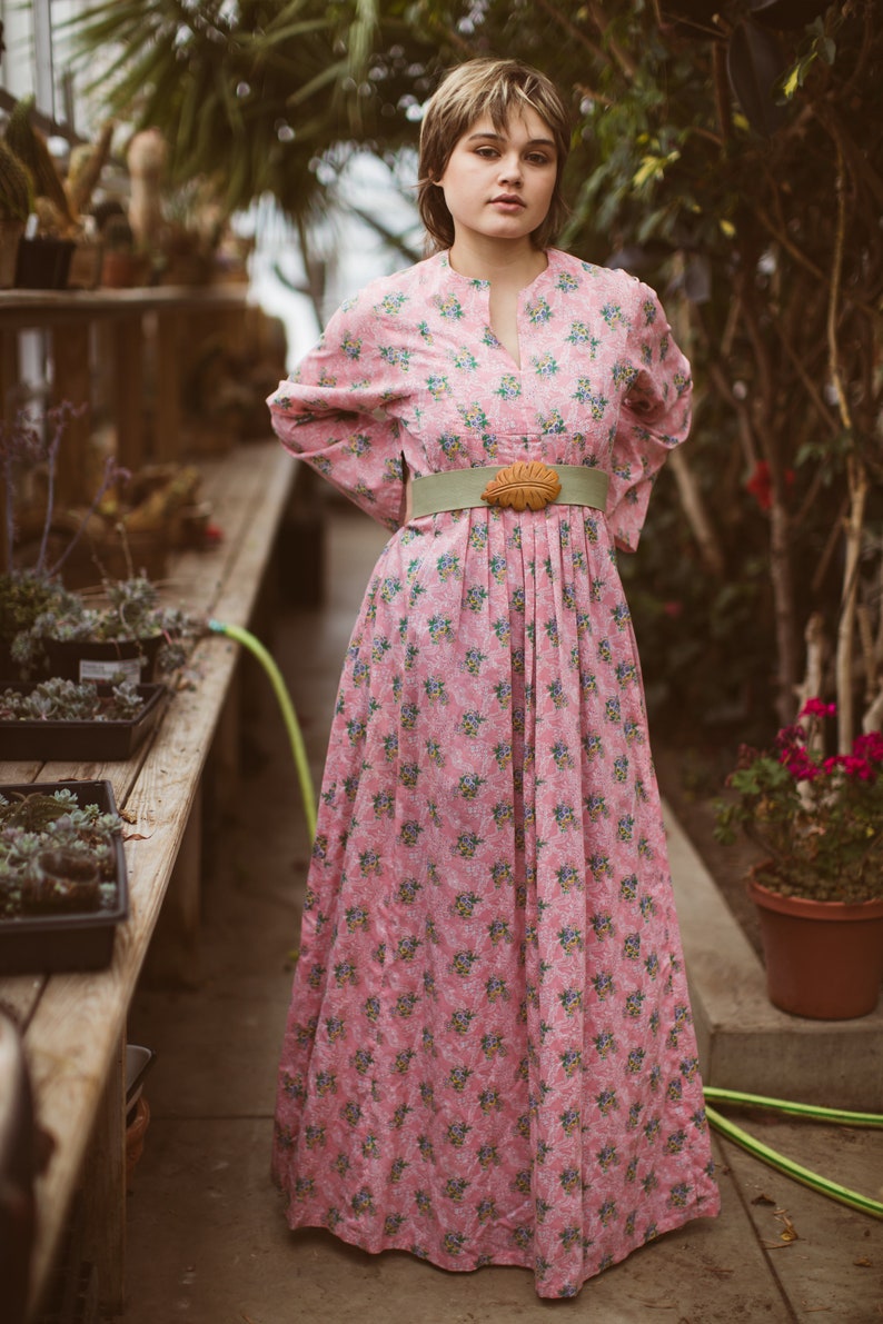 May include: A long pink dress with a floral pattern, featuring a high neckline and long sleeves. The dress is cinched at the waist with a light green belt and a decorative buckle. The dress is worn in a greenhouse setting.